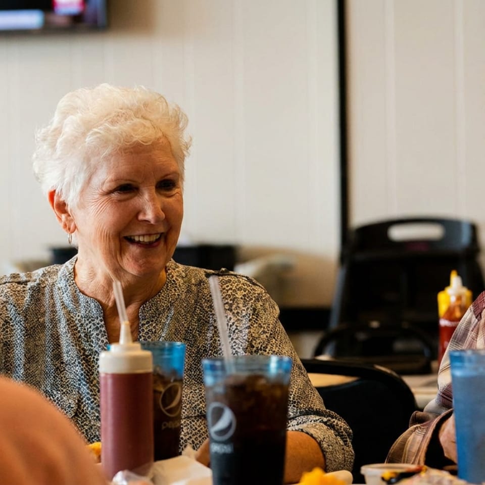 Smiling older woman enjoying a meal at a restaurant table with drinks and friends.