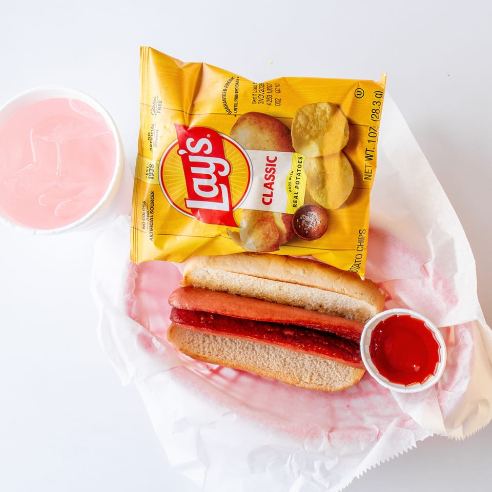 Hot dog in a bun with ketchup, a bag of Lay's potato chips, and a pink drink.