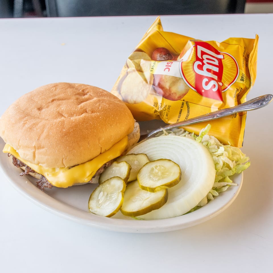 Cheeseburger with a side of pickles, onions, lettuce, and a bag of Lay's potato chips.