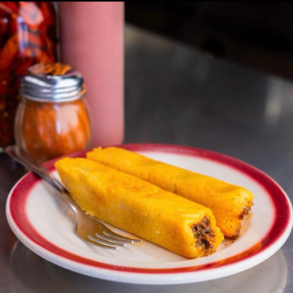 Two golden rolled corn tortillas filled with meat on a red-rimmed plate, accompanied by a jar of salsa and fork.