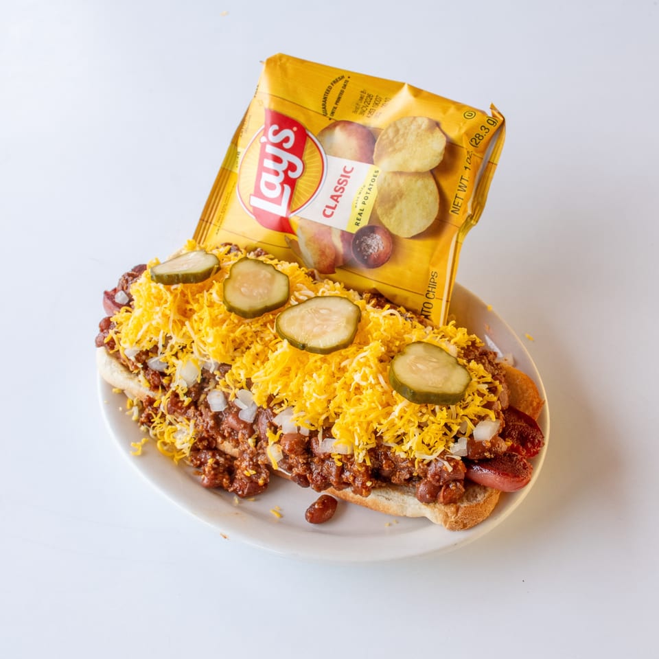 A chili dog with cheese, onions, and pickles, served with a bag of Lay's potato chips.