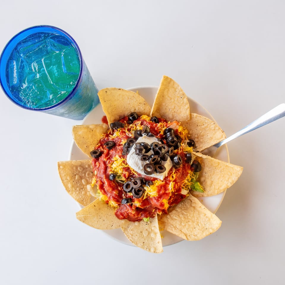 Overhead view of a taco salad with tortilla chips, salsa, cheese, olives, and sour cream, with a blue drink.